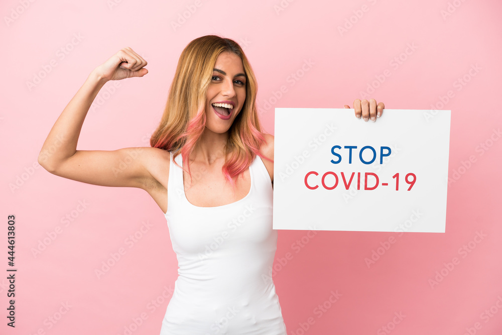 Young woman over isolated pink background holding a placard with text Stop Covid 19 and doing strong gesture Young woman over isolated pink background holding a placard with text Stop Covid 19 and doing strong gesture