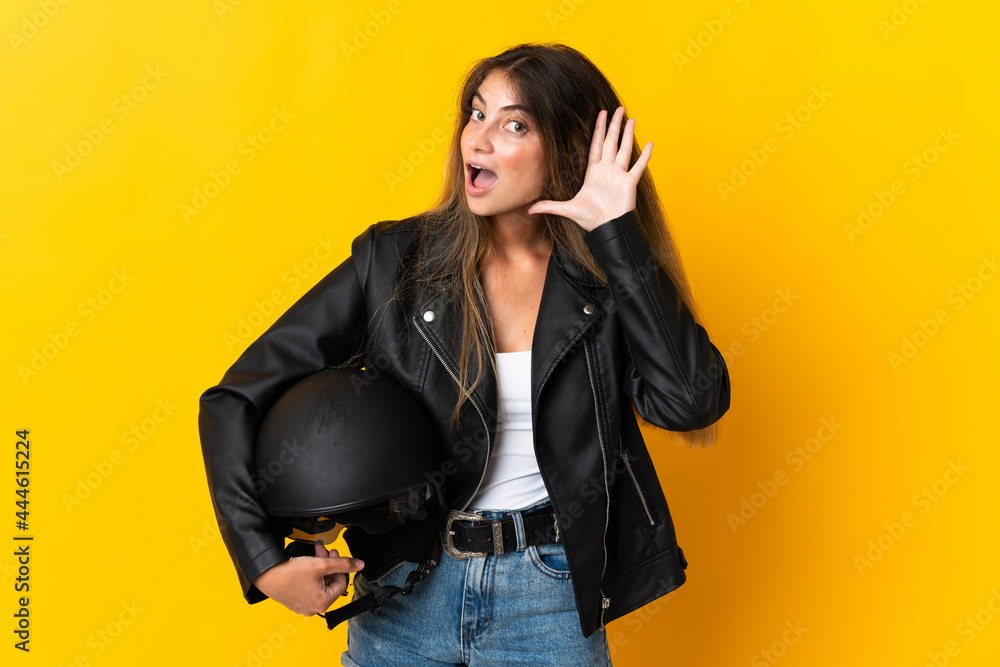 Woman holding a motorcycle helmet isolated on yellow background listening to something by putting hand on the ear Woman holding a motorcycle helmet isolated on yellow background listening to something by putting hand on the ear