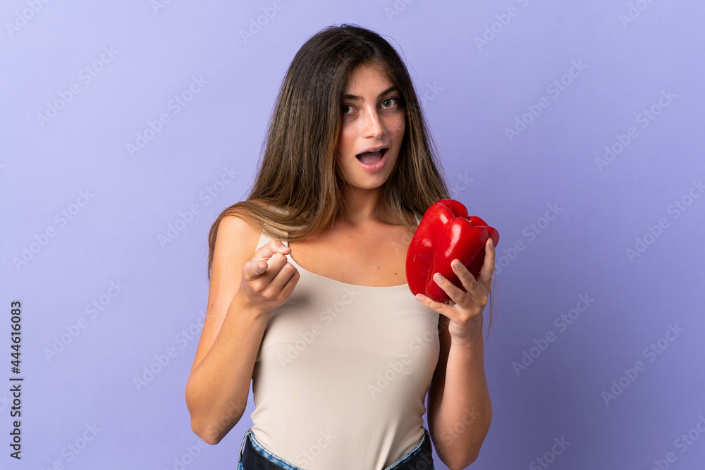Young woman holding a pepper isolated on purple background surprised and pointing front Young woman holding a pepper isolated on purple background surprised and pointing front