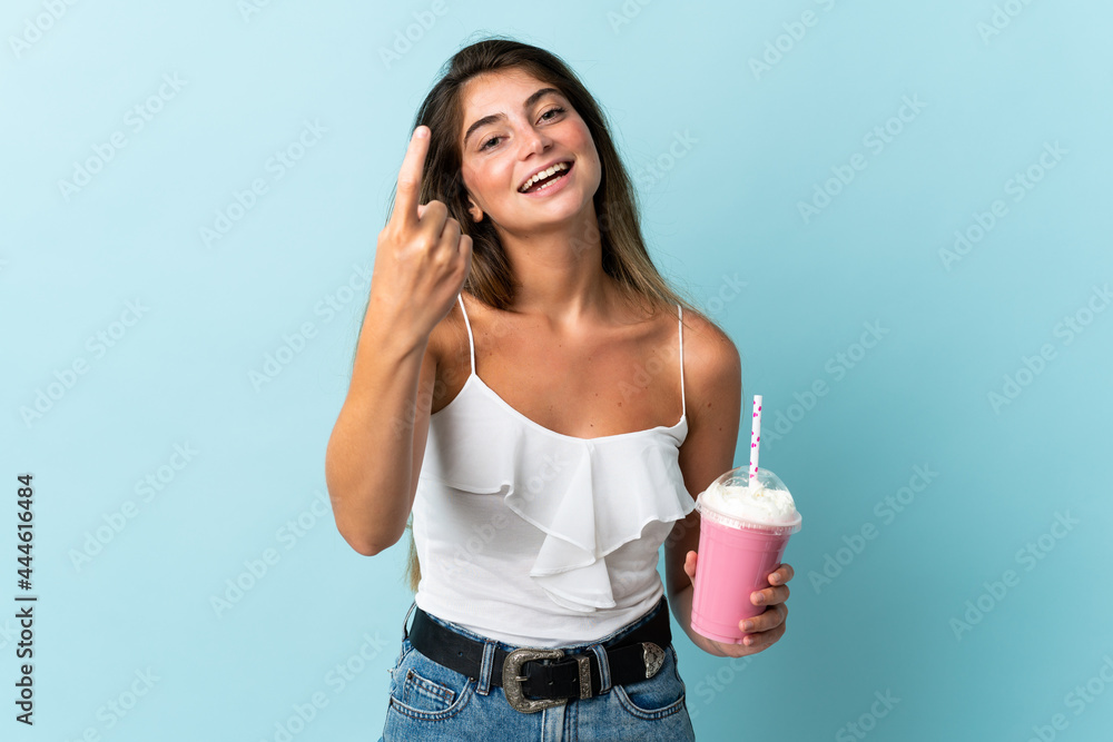Young woman with strawberry milkshake isolated on blue background doing coming gesture Young woman with strawberry milkshake isolated on blue background doing coming gesture