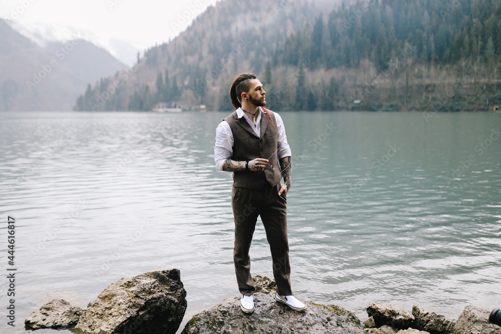 A brutal thoughtful young man groom with a dreadlocked hairstyle in a fashionable elegant wedding suit stands on the shore of a lake against the background of misty mountains in nature A brutal thoughtful young man groom with a dreadlocked hairstyle in a fashionable elegant wedding suit stands on the shore of a lake against the background of misty mountains in nature