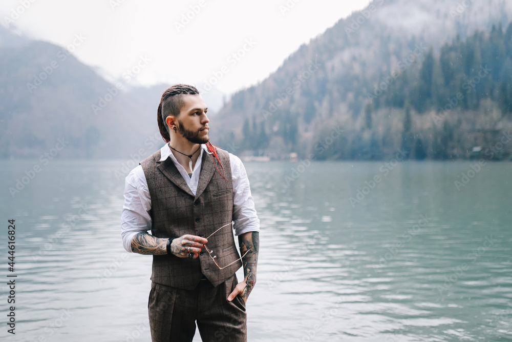 Fototapeta premium A brutal thoughtful young man groom with a dreadlocked hairstyle in a fashionable elegant wedding suit stands on the shore of a lake against the background of misty mountains in nature
