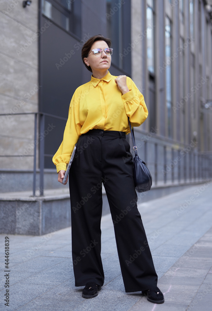 Attractive caucasian brunette businesswoman in black trousers and yellow blouse standing in front of office building. Urban background, finance or insurance concept. High quality vertical photo Attractive caucasian brunette businesswoman in black trousers and yellow blouse standing in front of office building. Urban background, finance or insurance concept. High quality vertical photo
