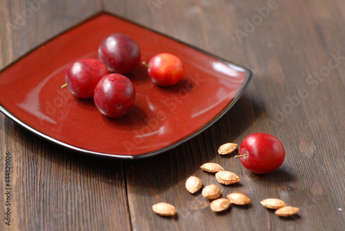 A red square dish with several fruits of red ripe cherry plums. Next to it on a wooden table there are some pips.