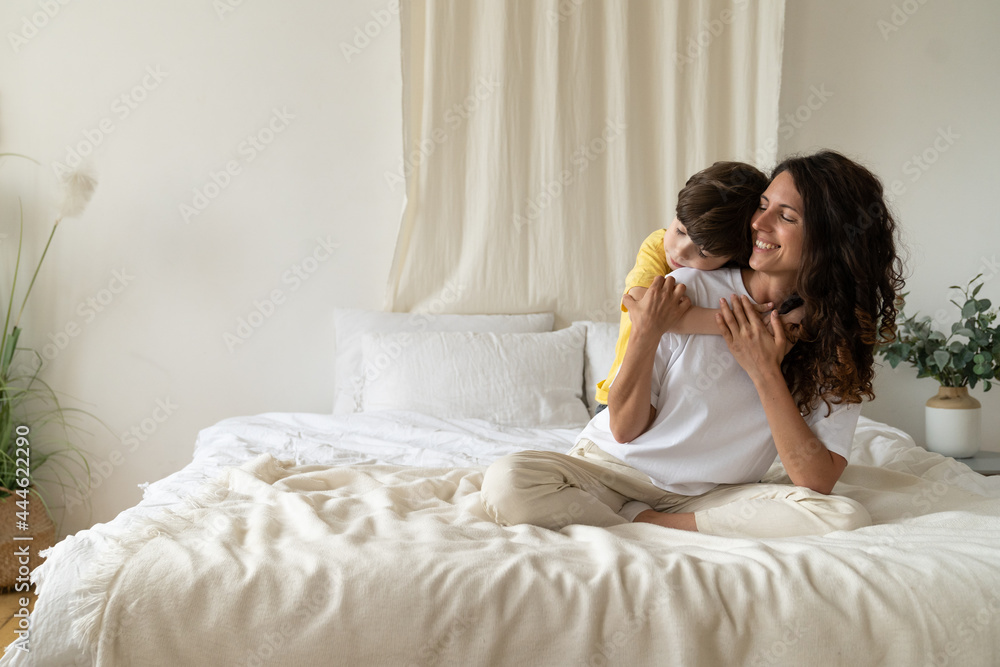 Cute kid boy hugging happy mom from back in morning sit on bed wearing pajamas. Caring mother enjoy tender time of closeness and togetherness with small child at home on weekend. Family love concept