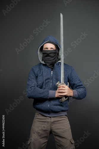 American Brutal man in a blue hoodie and black face mask with a Fantasy Handle Sword, stern look. Portrait of a courageous warrior in jeans and a hood holding a big sword with two hands after a battle