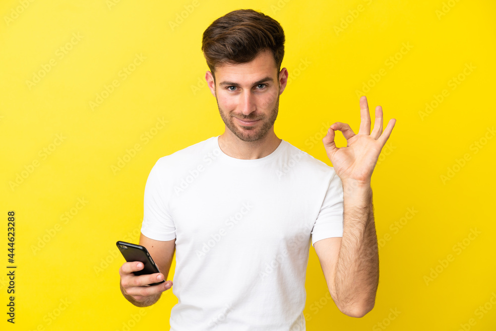 Young caucasian handsome man isolated on yellow background using mobile phone and doing OK sign Young caucasian handsome man isolated on yellow background using mobile phone and doing OK sign