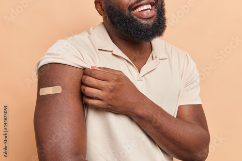 Cropped shot of unrecognizable man with thick beard shows arm with plaster glad to be vaccineted takes care of health dressed in casual t shirt isolated over brown background. Antivirus imunization photo