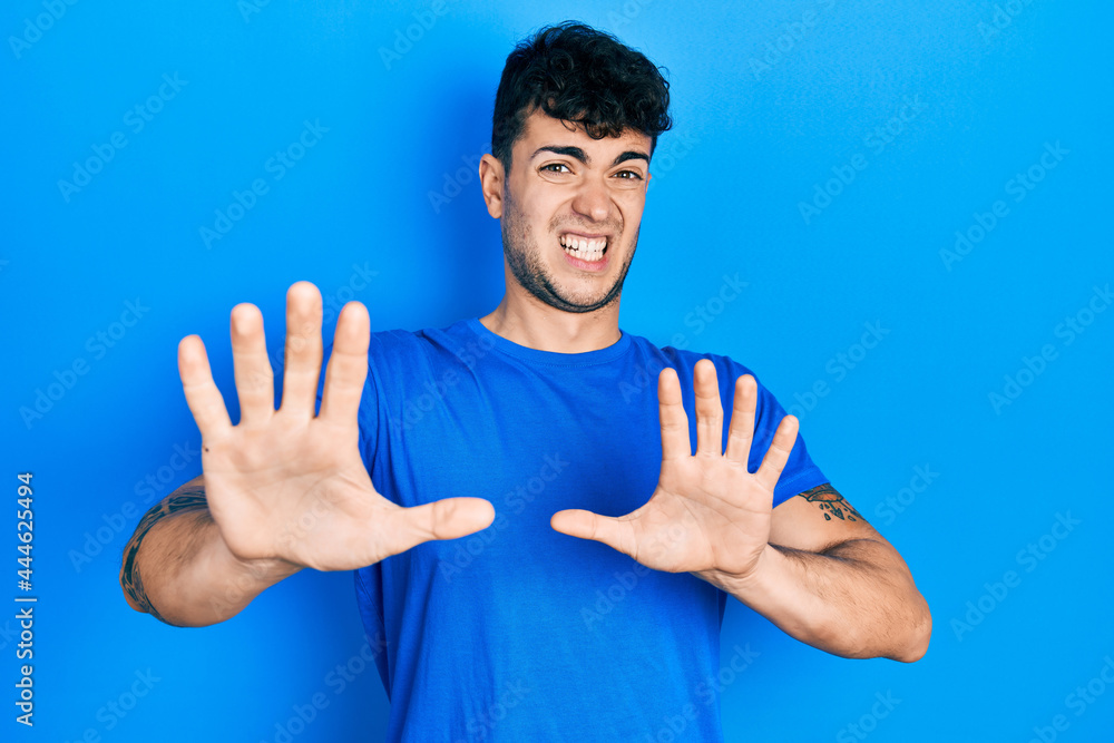 Young hispanic man wearing casual blue t shirt afraid and terrified with fear expression stop gesture with hands, shouting in shock. panic concept. Young hispanic man wearing casual blue t shirt afraid and terrified with fear expression stop gesture with hands, shouting in shock. panic concept.