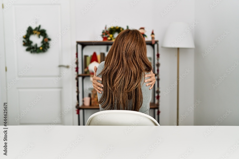 Young latin woman sitting on the table by christmas decor hugging oneself happy and positive from backwards. self love and self care Young latin woman sitting on the table by christmas decor hugging oneself happy and positive from backwards. self love and self care