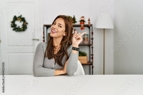 Young latin woman sitting on the table by christmas decor with a big smile on face, pointing with hand finger to the side looking at the camera.