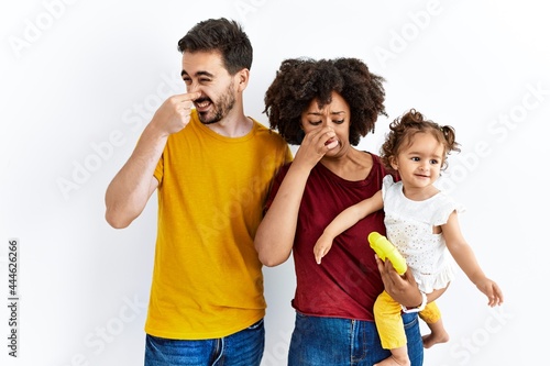 Interracial young family of black mother and hispanic father with daughter smelling something stinky and disgusting, intolerable smell, holding breath with fingers on nose. bad smell