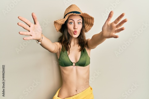 Young brunette woman wearing bikini and hat with open arms hugging making fish face with mouth and squinting eyes, crazy and comical.