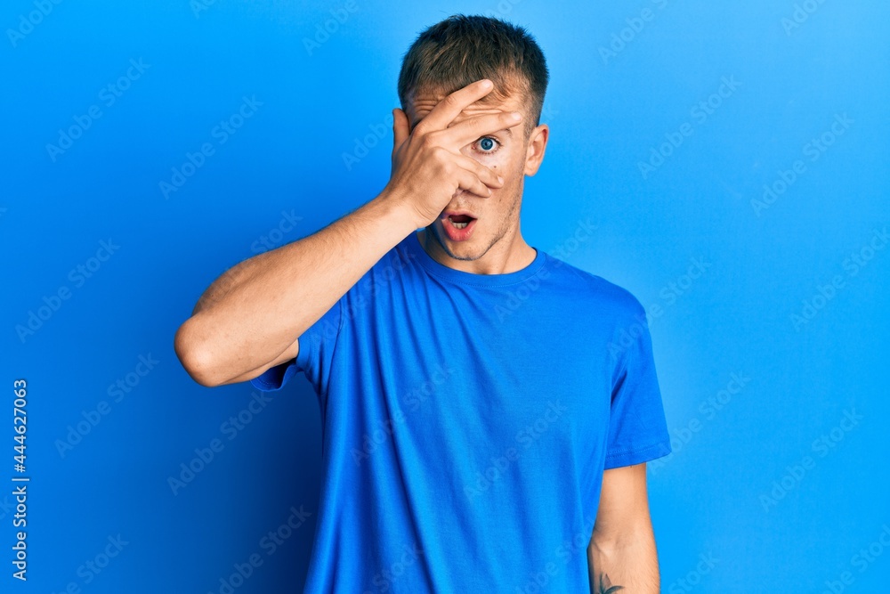 Young caucasian man wearing casual blue t shirt peeking in shock covering face and eyes with hand, looking through fingers with embarrassed expression. Young caucasian man wearing casual blue t shirt peeking in shock covering face and eyes with hand, looking through fingers with embarrassed expression.