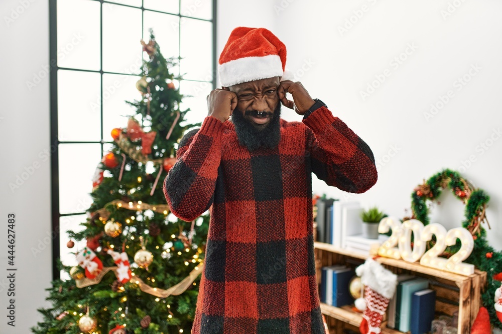 African american man wearing santa claus hat standing by christmas tree covering ears with fingers with annoyed expression for the noise of loud music. deaf concept.