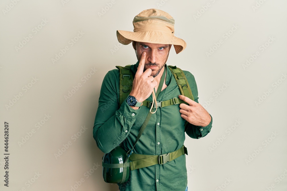Handsome man with beard wearing explorer hat and backpack pointing to the eye watching you gesture, suspicious expression Handsome man with beard wearing explorer hat and backpack pointing to the eye watching you gesture, suspicious expression