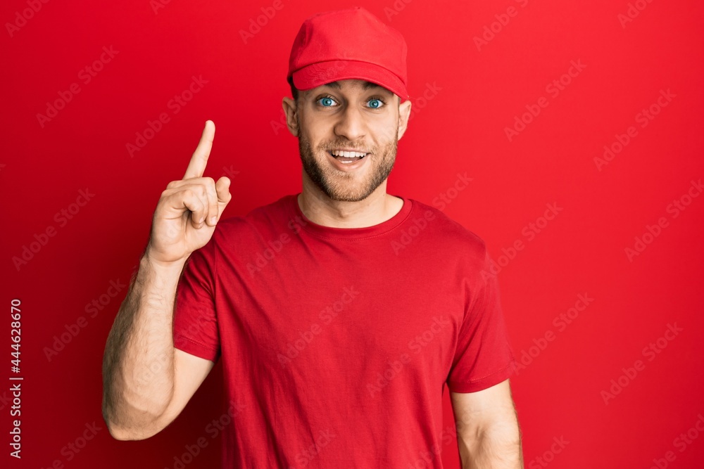 Young caucasian man wearing delivery uniform and cap pointing finger up with successful idea. exited and happy. number one. Young caucasian man wearing delivery uniform and cap pointing finger up with successful idea. exited and happy. number one.