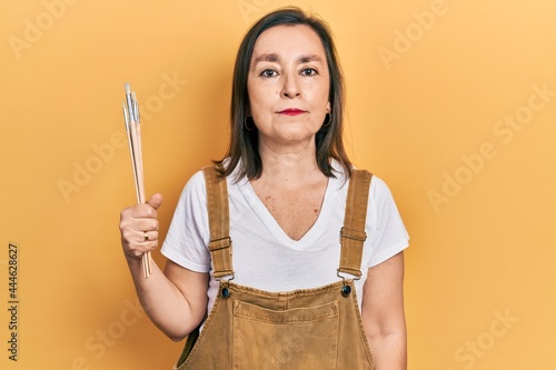 Middle age hispanic woman holding paintbrushes thinking attitude and sober expression looking self confident