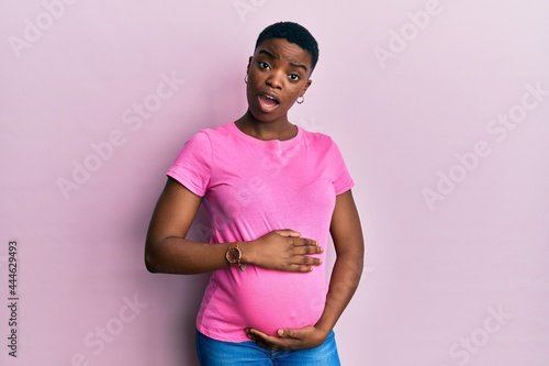 Young african american woman expecting a baby, touching pregnant belly in shock face, looking skeptical and sarcastic, surprised with open mouth