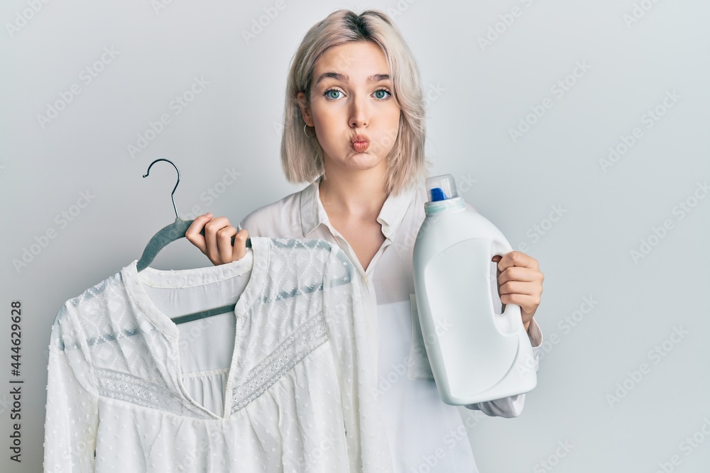 Young blonde girl holding detergent bottle and shirt puffing cheeks with funny face. mouth inflated with air, catching air. Young blonde girl holding detergent bottle and shirt puffing cheeks with funny face. mouth inflated with air, catching air.