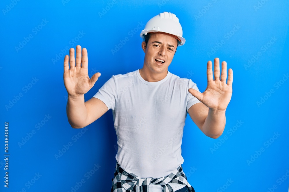 Handsome young man wearing builder uniform and hardhat afraid and terrified with fear expression stop gesture with hands, shouting in shock. panic concept.