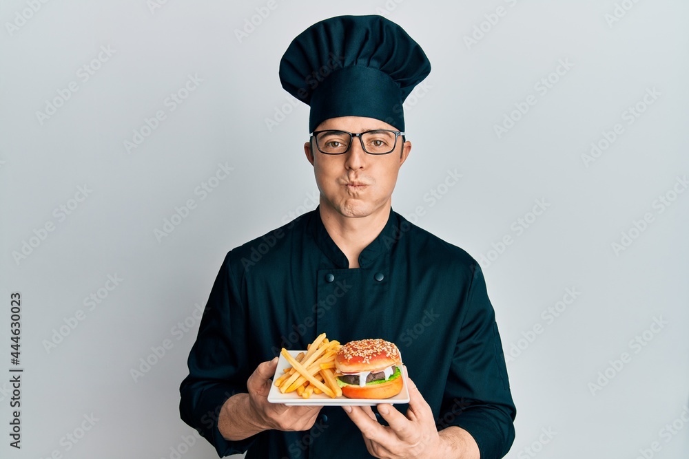 Handsome young man chef holding burger with fries puffing cheeks with funny face. mouth inflated with air, catching air. Handsome young man chef holding burger with fries puffing cheeks with funny face. mouth inflated with air, catching air.