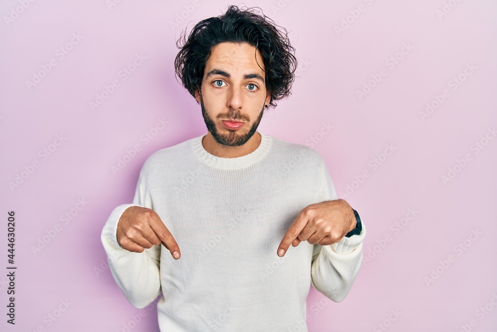 Handsome hispanic man wearing casual white sweater pointing down looking sad and upset, indicating direction with fingers, unhappy and depressed. Handsome hispanic man wearing casual white sweater pointing down looking sad and upset, indicating direction with fingers, unhappy and depressed.