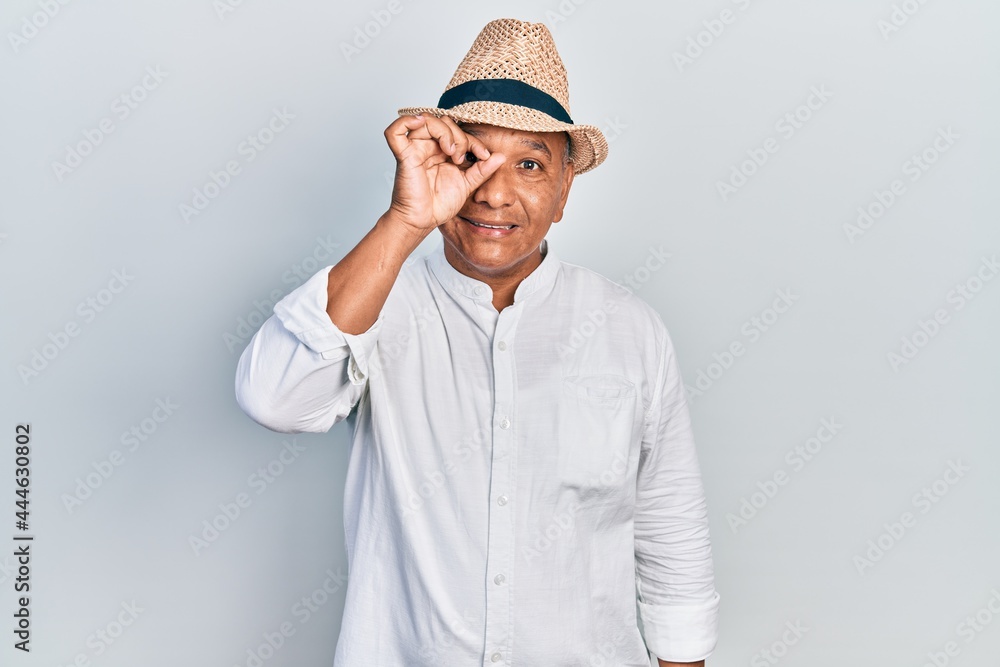 Middle age latin man wearing casual clothes and hat smiling happy doing ok sign with hand on eye looking through fingers Middle age latin man wearing casual clothes and hat smiling happy doing ok sign with hand on eye looking through fingers