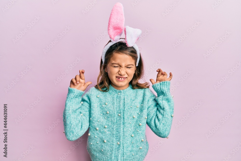 Little beautiful girl wearing cute easter bunny ears gesturing finger crossed smiling with hope and eyes closed. luck and superstitious concept. Little beautiful girl wearing cute easter bunny ears gesturing finger crossed smiling with hope and eyes closed. luck and superstitious concept.