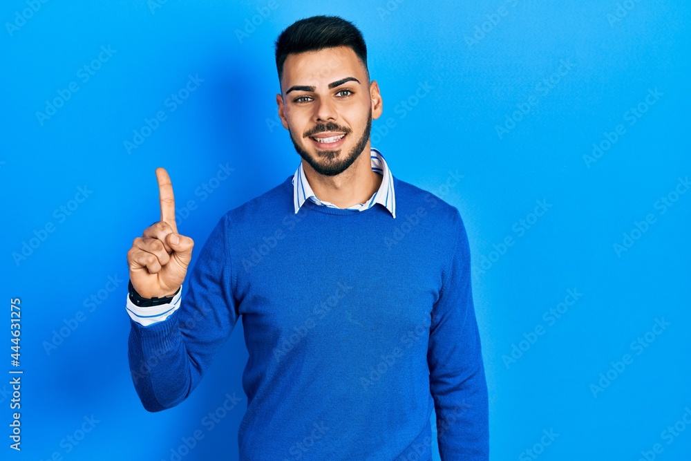 Fototapeta premium Young hispanic man with beard wearing casual blue sweater pointing finger up with successful idea. exited and happy. number one.