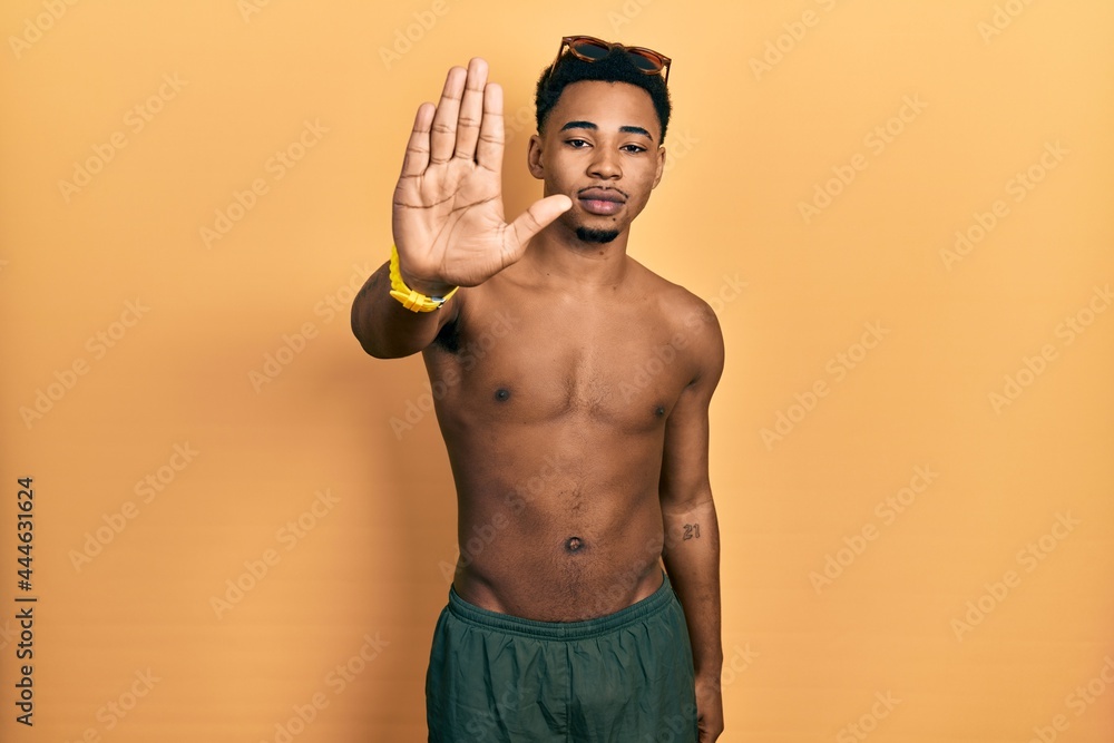 Young african american man wearing swimwear and swimmer glasses doing stop sing with palm of the hand. warning expression with negative and serious gesture on the face.