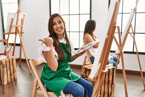 Young hispanic artist women painting on canvas at art studio doing ok gesture with hand smiling, eye looking through fingers with happy face.