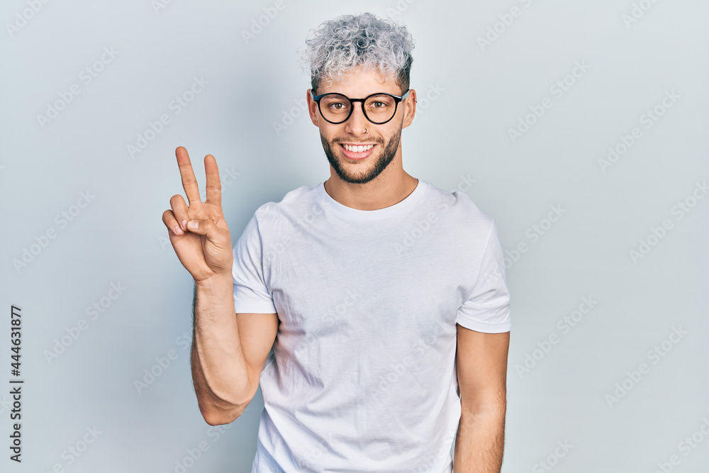 Young hispanic man with modern dyed hair wearing white t shirt and glasses smiling looking to the camera showing fingers doing victory sign. number two. Young hispanic man with modern dyed hair wearing white t shirt and glasses smiling looking to the camera showing fingers doing victory sign. number two.