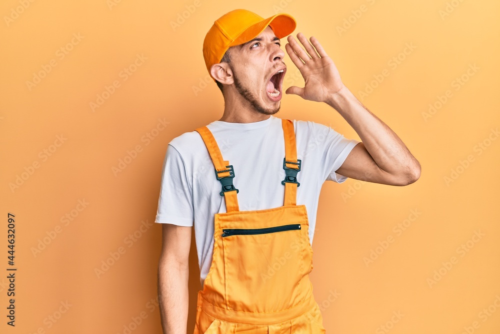 Hispanic young man wearing handyman uniform shouting and screaming loud to side with hand on mouth. communication concept. Hispanic young man wearing handyman uniform shouting and screaming loud to side with hand on mouth. communication concept.