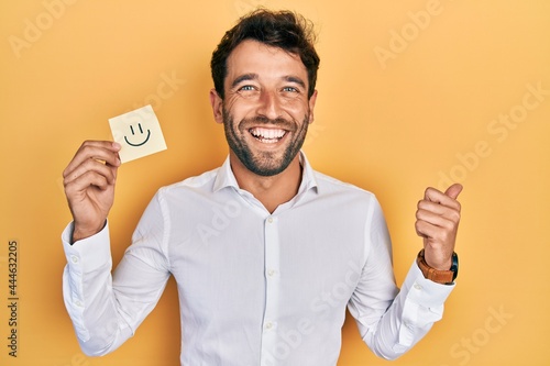 Handsome man with beard holding smile emoji reminder pointing thumb up to the side smiling happy with open mouth © Krakenimages.com