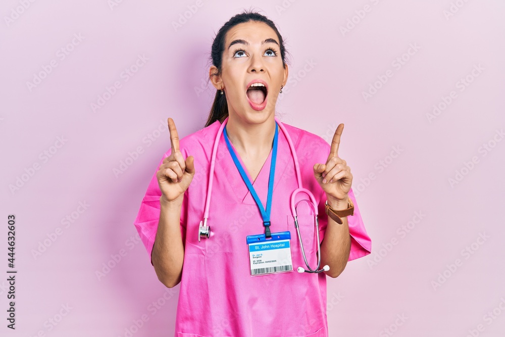 Young brunette woman wearing doctor uniform and stethoscope amazed and surprised looking up and pointing with fingers and raised arms. Young brunette woman wearing doctor uniform and stethoscope amazed and surprised looking up and pointing with fingers and raised arms.