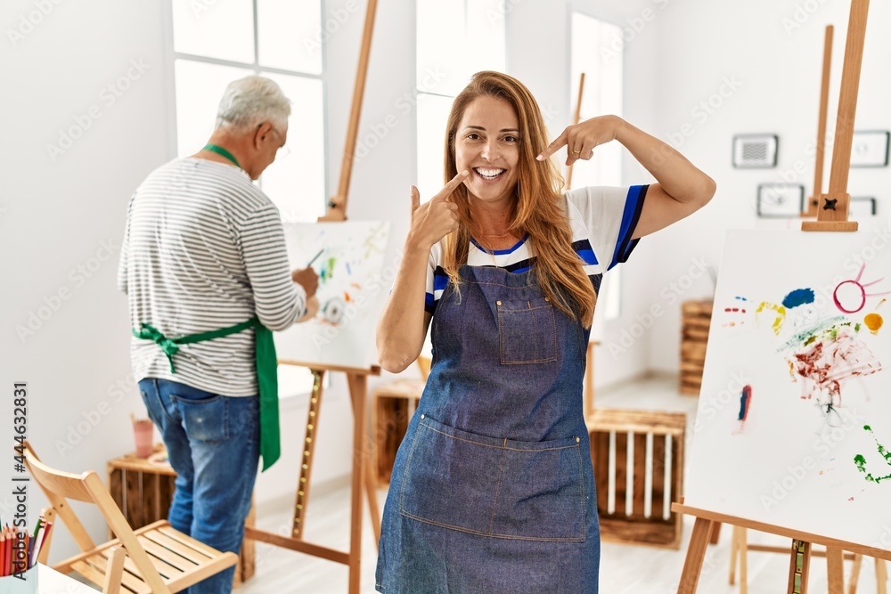 Hispanic woman wearing apron at art studio smiling cheerful showing and pointing with fingers teeth and mouth. dental health concept.