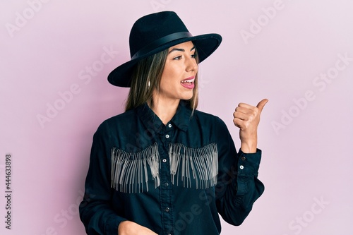 Young hispanic woman wearing cowgirl style smiling with happy face looking and pointing to the side with thumb up.