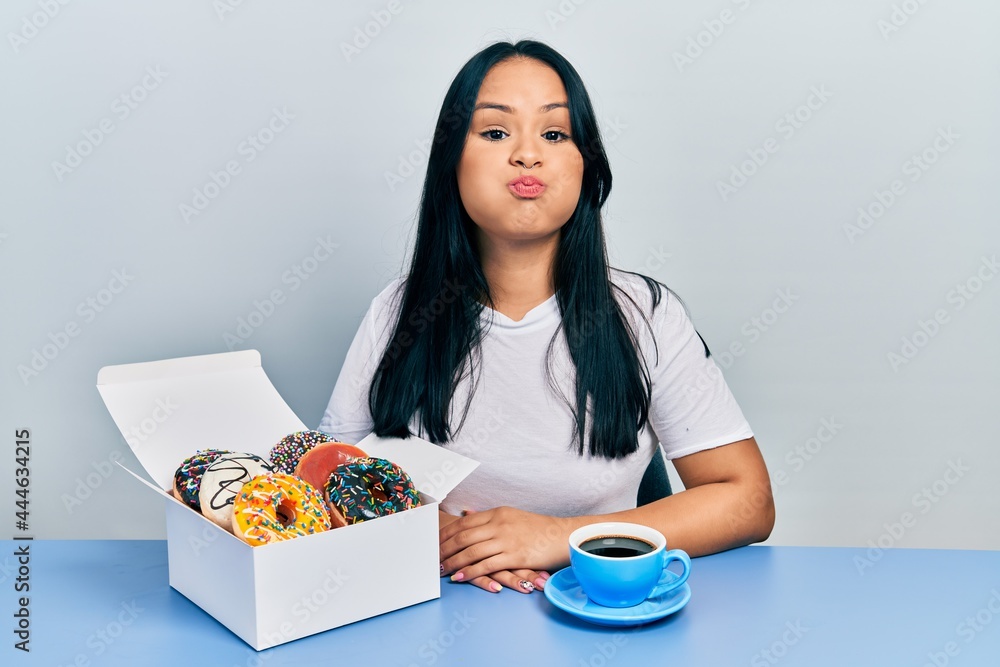 Beautiful hispanic woman with nose piercing eating doughnuts and drinking a cup of coffee puffing cheeks with funny face. mouth inflated with air, catching air. Beautiful hispanic woman with nose piercing eating doughnuts and drinking a cup of coffee puffing cheeks with funny face. mouth inflated with air, catching air.
