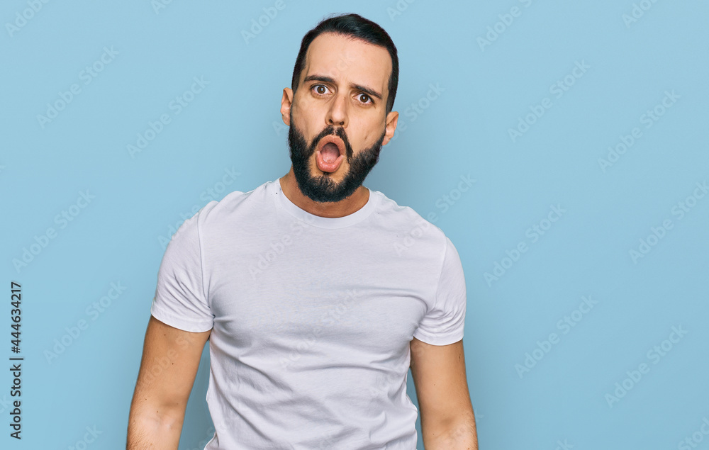 Young man with beard wearing casual white t shirt in shock face, looking skeptical and sarcastic, surprised with open mouth Young man with beard wearing casual white t shirt in shock face, looking skeptical and sarcastic, surprised with open mouth