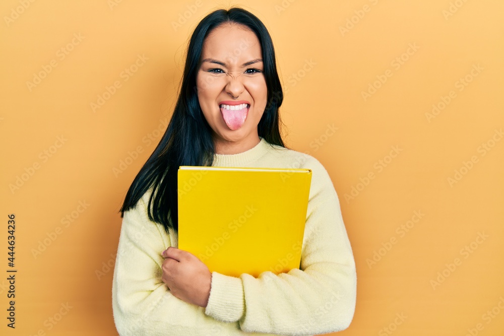Beautiful hispanic woman with nose piercing holding book sticking tongue out happy with funny expression. Beautiful hispanic woman with nose piercing holding book sticking tongue out happy with funny expression.