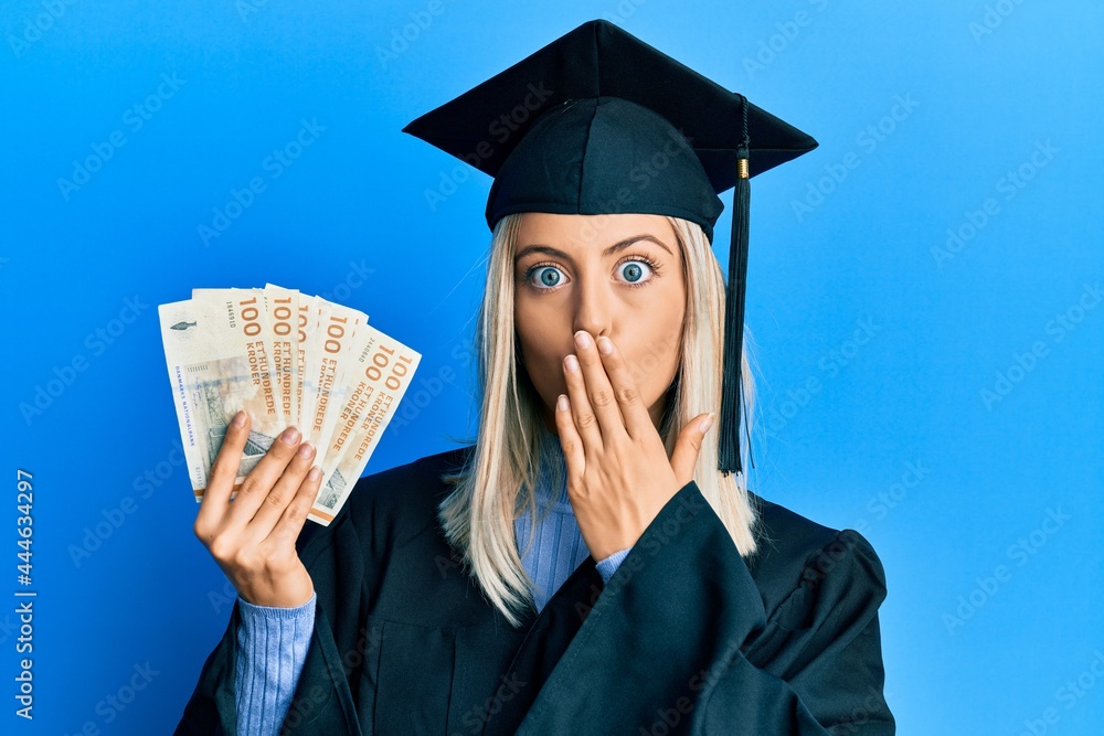 Beautiful blonde woman wearing graduation cap and ceremony robe holding denmark krone covering mouth with hand, shocked and afraid for mistake. surprised expression Beautiful blonde woman wearing graduation cap and ceremony robe holding denmark krone covering mouth with hand, shocked and afraid for mistake. surprised expression