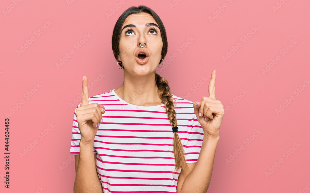 Young hispanic woman wearing casual clothes amazed and surprised looking up and pointing with fingers and raised arms. Young hispanic woman wearing casual clothes amazed and surprised looking up and pointing with fingers and raised arms.