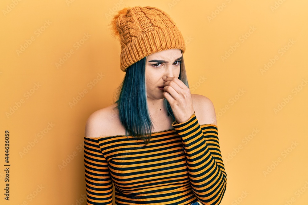 Young modern girl wearing wool hat smelling something stinky and disgusting, intolerable smell, holding breath with fingers on nose. bad smell Young modern girl wearing wool hat smelling something stinky and disgusting, intolerable smell, holding breath with fingers on nose. bad smell