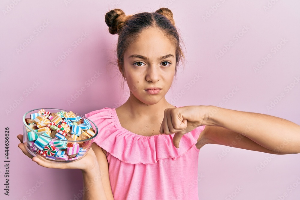 Beautiful brunette little girl holding jar full of sugar candies with angry face, negative sign showing dislike with thumbs down, rejection concept Beautiful brunette little girl holding jar full of sugar candies with angry face, negative sign showing dislike with thumbs down, rejection concept
