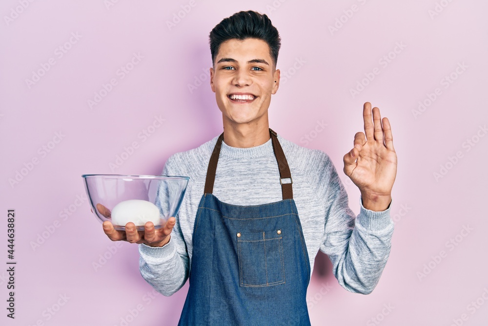 Young hispanic man wearing apron holding bowl of bread dough doing ok sign with fingers, smiling friendly gesturing excellent symbol Young hispanic man wearing apron holding bowl of bread dough doing ok sign with fingers, smiling friendly gesturing excellent symbol