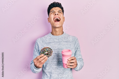 Young hispanic man eating doughnut and drinking coffee angry and mad screaming frustrated and furious, shouting with anger looking up.