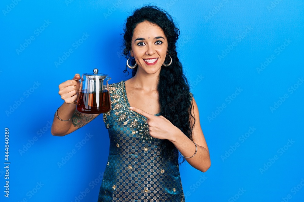 Young woman wearing bindi and traditional kurta holding traditional tea pot smiling happy pointing with hand and finger Young woman wearing bindi and traditional kurta holding traditional tea pot smiling happy pointing with hand and finger