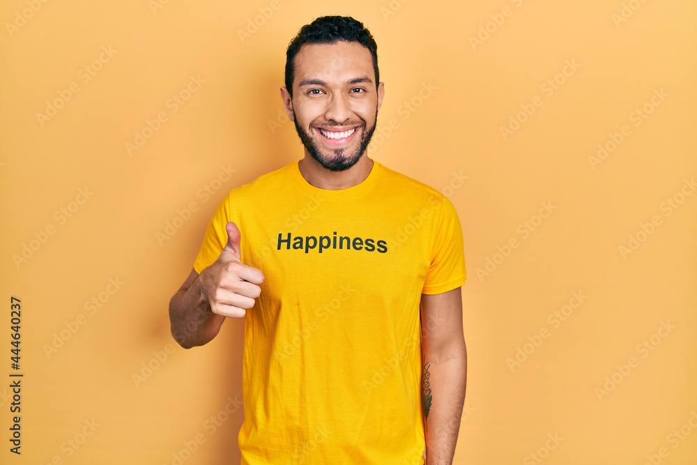 Hispanic man with beard wearing t shirt with happiness word message doing happy thumbs up gesture with hand. approving expression looking at the camera showing success. Hispanic man with beard wearing t shirt with happiness word message doing happy thumbs up gesture with hand. approving expression looking at the camera showing success.