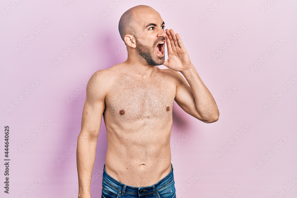 Young bald man standing shirtless shouting and screaming loud to side with hand on mouth. communication concept. Young bald man standing shirtless shouting and screaming loud to side with hand on mouth. communication concept.